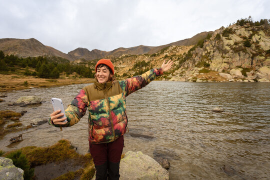 Woman in nature wearing winter hat, hiking in middle of mountains with lake in background looking at happy camera, making photos with the mobile phone making a call