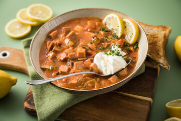 Ost Deutsche DDR GDR Soljanka Eintopf Suppe mit Jagdwurst, Saure Gurken, Letscho, Schmand, Zitrone, Toast und Petersilie mit Löffel, Holzbrett und grün Hintergrund