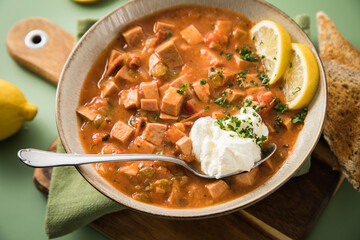 Ost Deutsche DDR GDR Soljanka Eintopf Suppe mit Jagdwurst, Saure Gurken, Letscho, Schmand, Zitrone, Toast und Petersilie mit Löffel, Holzbrett und grün Hintergrund