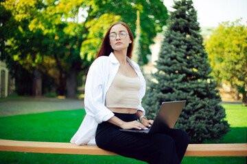 A female student in glasses sits on a bench near the university with a laptop. Freelance work in the fresh air