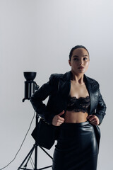 Young pretty woman in black lacy bra and leather suit posing near spotlight.