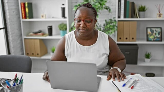 Confident plus size african american woman boss, working online and relaxed in the office interior, casting an elegant look around, focused on her laptop.