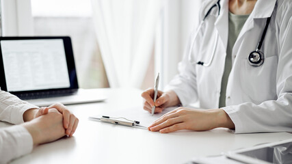 Doctor and patient sitting near each other at the white desk in clinic. Female physician is listening filling up a records form. Medicine concept