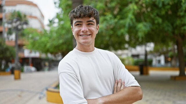 Cheerful young hispanic teenager, beaming with confidence, expressing joy through a playful arm crossed gesture in the heart of a lush green city park