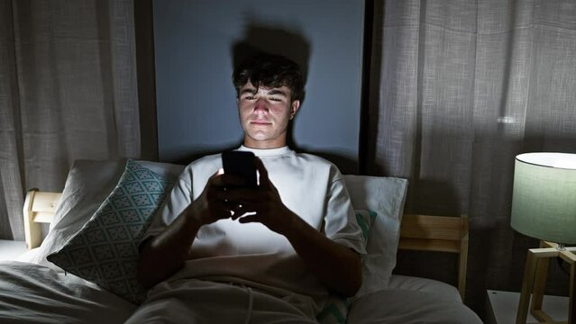 Chilled young hispanic teenager grinning wide, tapping at his phone, comfortably stretched out on his room's bed, enjoying the quiet night