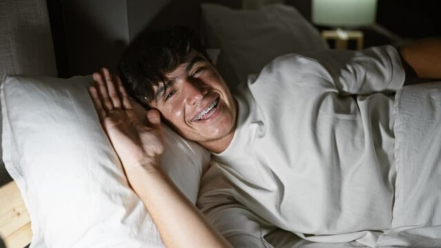 Handsome young hispanic teenager, with buoyant confidence, expressively enjoying his morning, reclining on a comfortable bed, amid the soft light in his cozy bedroom.