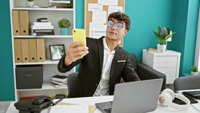 Handsome young hispanic teen worker in office papped snapping a selfie on his laptop while bossing business on his phone.