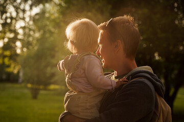 Father plays with a cute blonde baby toddler in the park at sunset. The concept of a happy childhood, a strong family. Sun rays with selective focus - obrazy, fototapety, plakaty