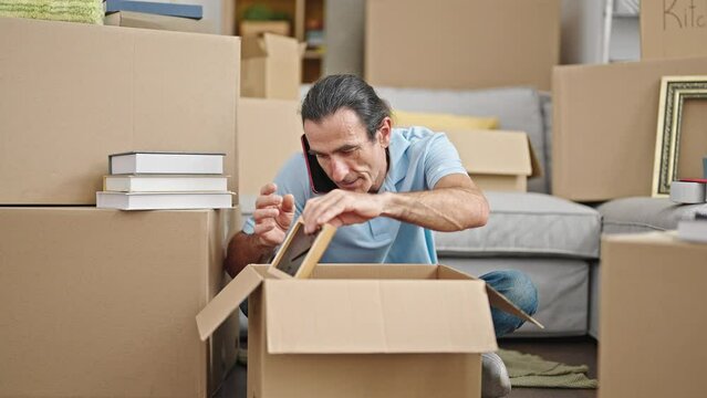 Middle age man packing books on cardboard box talking on smartphone at new home