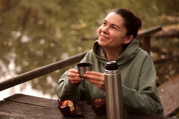 young woman smiling drinking tea from thermos with muffin outdoors