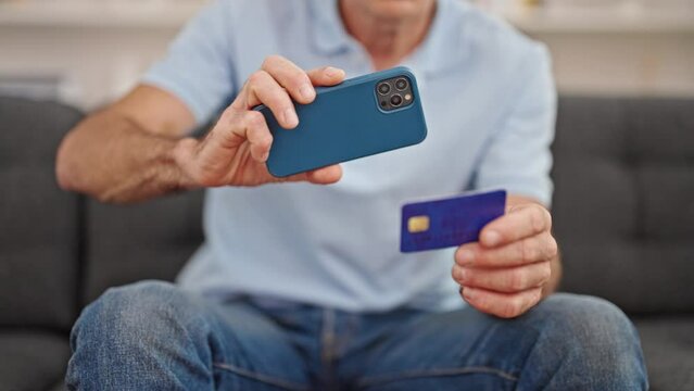 Middle age man taking photo to credit card by smartphone sitting on sofa at home