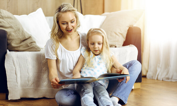 Happy family. Blonde young mother reading a book to her cute daughter while sitting at wooden floor in sunny room. Motherhood concept