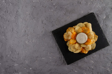 Top view of heap of potato chips with creamy white dipping sauce on black slate serving board on...