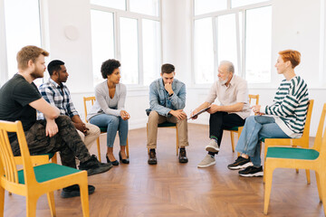 Wide shot of bearded aged professional male mentor leader counselor coach speaking at diverse business meeting with team people or patients group, during training psychotherapy session. - obrazy, fototapety, plakaty