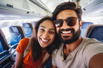 Fotobehang Happy indian tourist couple taking a selfie inside an airplane. Positive young couple on a vacation taking a selfie in a plane before takeoff. © Katrin Kovac