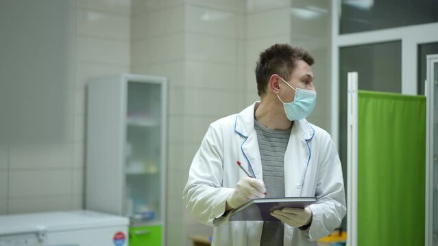 A male medical worker, wearing a mask and a white coat, nervously writes something on the cover of a medical journal