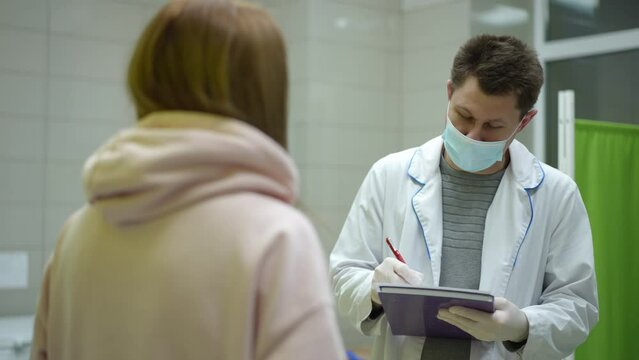 A male medical worker, wearing a white coat, mask and rubber gloves, filled a questionnaire with a pen, according to the patient