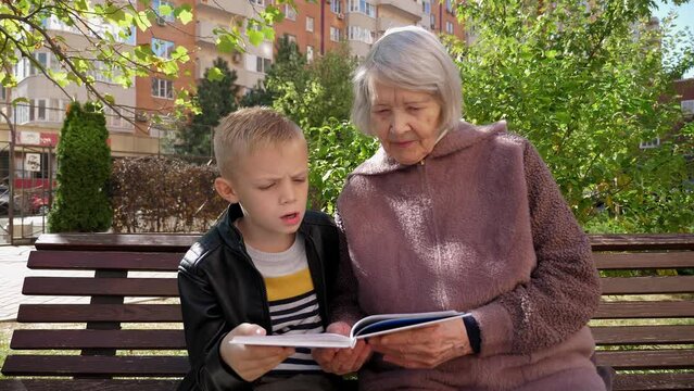 An elderly woman with gray hair and her grandson are reading a book together in the park on a bench, the grandson spends time with an old grandmother. Stable image.