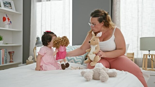 Confident mother and cheerful daughter enjoying playtime with toys, sitting on a comfy bed in a sunlit bedroom, expressing joy and love with warm smiles