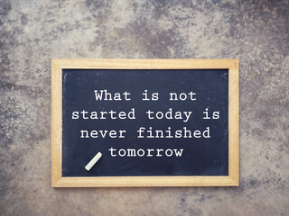 Motivational and inspirational wording. What Is Not Started Today Is Never Finished Tomorrow written on a blackboard. With blurred styled background.