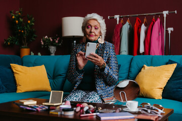 Confident elderly gray hair woman in fashionable wear using smart phone while sitting on the couch 