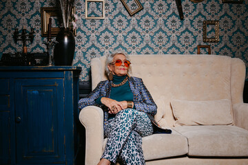 Stylish senior woman in trendy and luxury wear sitting on comfortable couch at retro styled interior