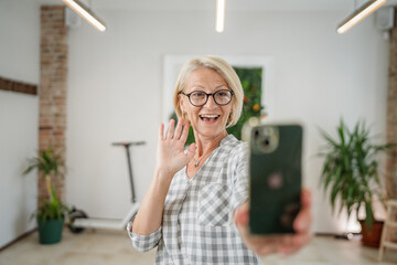 One woman mature caucasian female at home or office use mobile phone hold smartphone take selfie photos self portrait or make a video call daily routine copy space