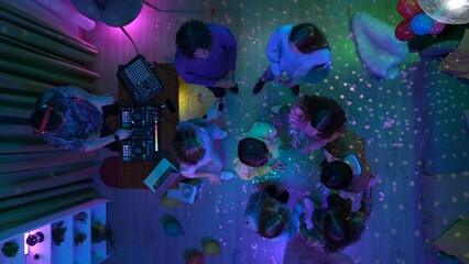Group of teenagers dancing in a decorated party room with a disco ball. A man is mixing music on a dj console. Neon colorful lights. Top view.