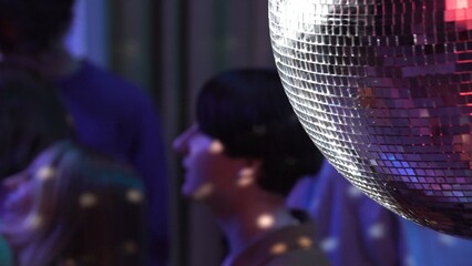 Close up shot capturing a glowing disco ball. Blurred picture of a group of teenagers dancing in a...