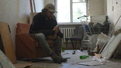 Homeless poor man sitting in a room of an abandoned building filled with his meager belongings. He is holding a bottle of alcohol in his hand, looking frustrated.