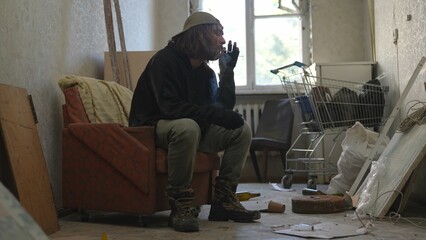 Homeless poor man sitting in a room of an abandoned building filled with his meager belongings. He...