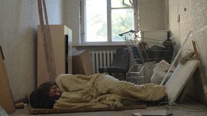 Homeless poor man sleeping on a piece of cardboard, covered with a blanket in a room of an...