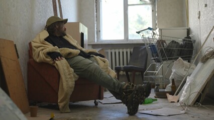 Homeless poor man covered in a blanket sitting in a room of an abandoned building filled with his...