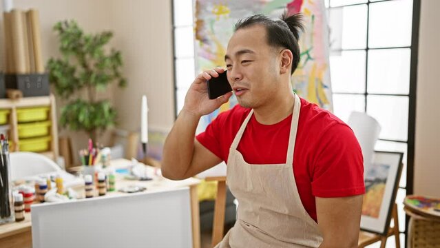 Confident young chinese artist, cheerfully talking on smartphone in his art studio, filled with canvas and vibrant paint palette