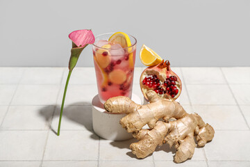 Glass of tasty pomegranate cocktail with lemon, ginger root and calla lily on white tile near grey wall