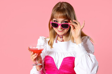 Obraz premium Young woman dressed as doll with cocktail on pink background