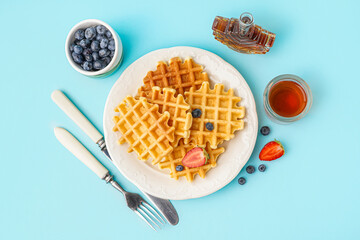 Plate of tasty waffles with berries and maple syrup on blue background