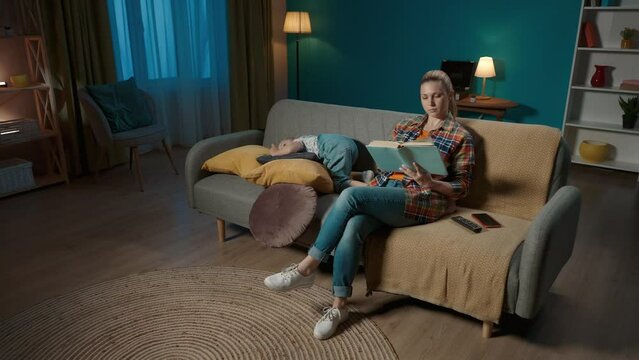 A little girl is rummaging around on the couch in the cushions, jumping around while her mom is reading a book and not paying attention to her. Mom and daughter sitting on the couch in the living room