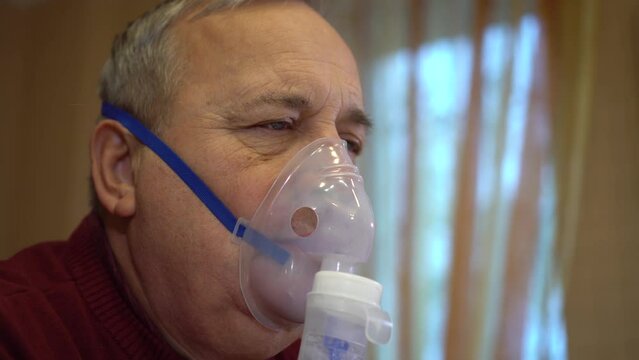 An elderly man inhales through an oxygen mask. A man does a respiratory procedure at home. Closeup.