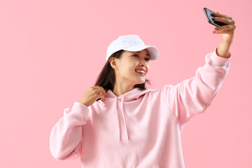 Beautiful young Asian woman in stylish hoodie taking selfie on pink background