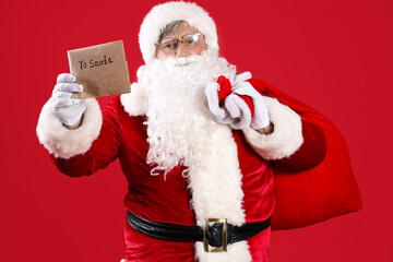 Santa Claus with letter and bag full of Christmas gifts on red background