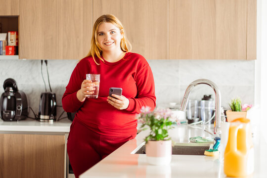 Smiling young plus size woman holding glass of water, using mobile phone, looking at camera.