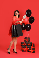 Beautiful African-American woman with balloons, gift boxes, shopping bags and money on red background. Black Friday sale