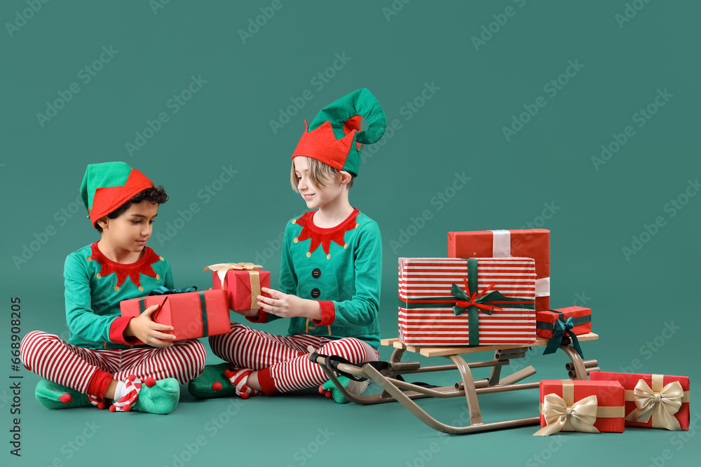 Sticker Cute little children in elves costumes with Christmas gifts and sledge on green background