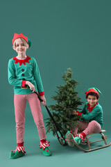 Cute little children in elves costumes with Christmas tree and sledge on green background
