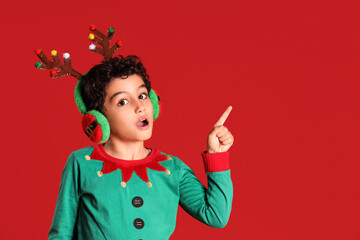Cute little boy in elf costume with deer horns pointing at something on red background