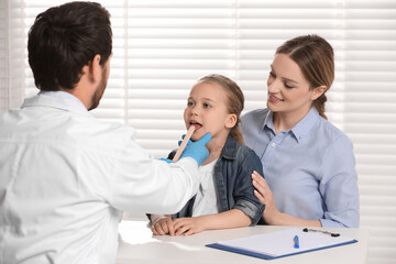 Fototapeta premium Doctor examining girl`s oral cavity with tongue depressor near her mother indoors