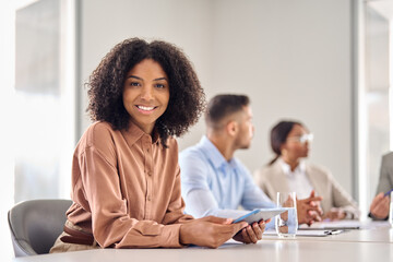 Happy African American company employee, smiling young business woman sitting at diverse team office meeting. Professional businesswoman company employee leader with tablet in board room, portrait. - obrazy, fototapety, plakaty