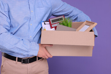 Unemployed man with box of personal office belongings on purple background, closeup