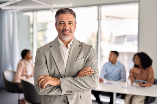 Confident older middle aged Indian business man manager standing at office team meeting. Portrait of elegant smiling professional senior male company executive leader crossing arms in board room.